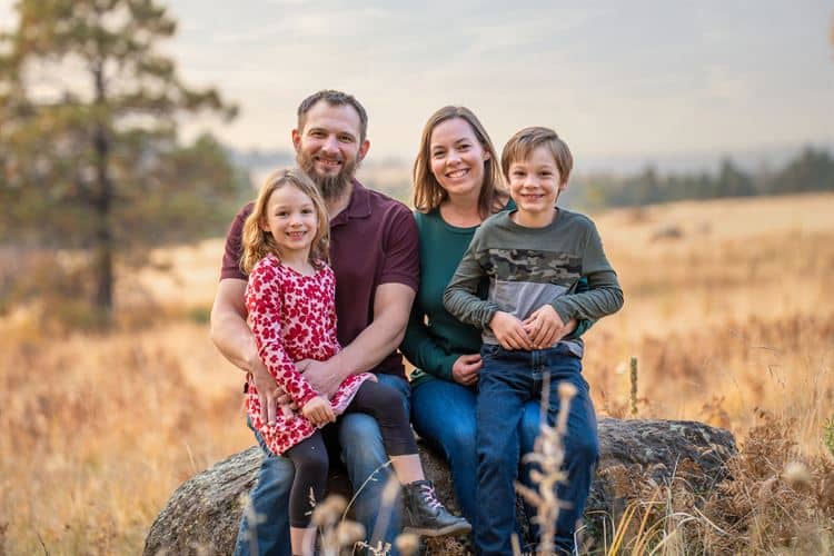 A family of four smiling and sitting on a large rock in a grassy field, with trees and a blurred natural background. A family of four smiling and sitting on a large rock in a grassy field, with trees and a blurred natural background.